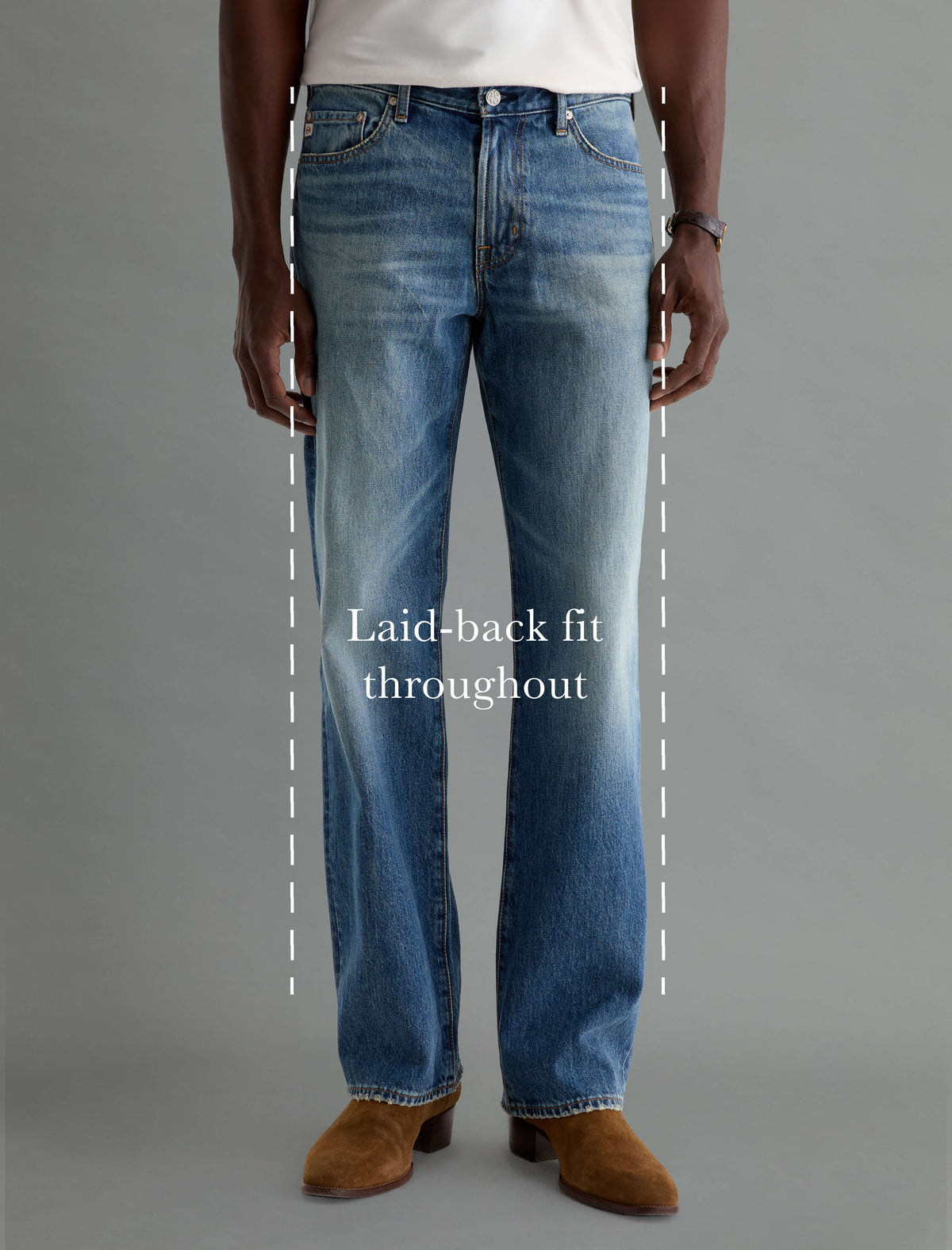 A model wearing medium-wash blue jeans with a straight-leg or laid-back fit, shown from the waist down against a plain gray studio background. White dashed lines and the text 'Laid-back fit throughout' highlight the loose silhouette of the jeans.