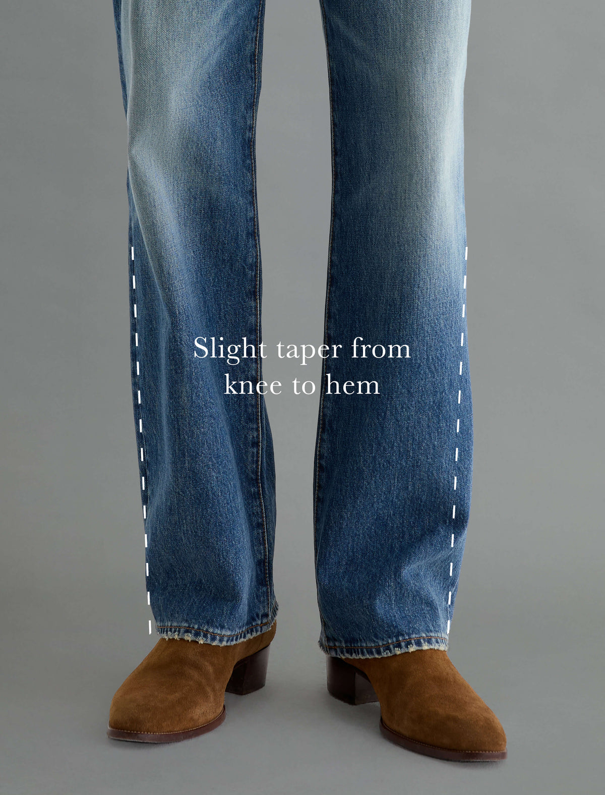 Close-up of a person's legs wearing blue jeans and brown suede ankle boots against a plain gray studio background, with white text reading 'Slight taper from knee to hem' and dashed line graphics indicating the leg shape.