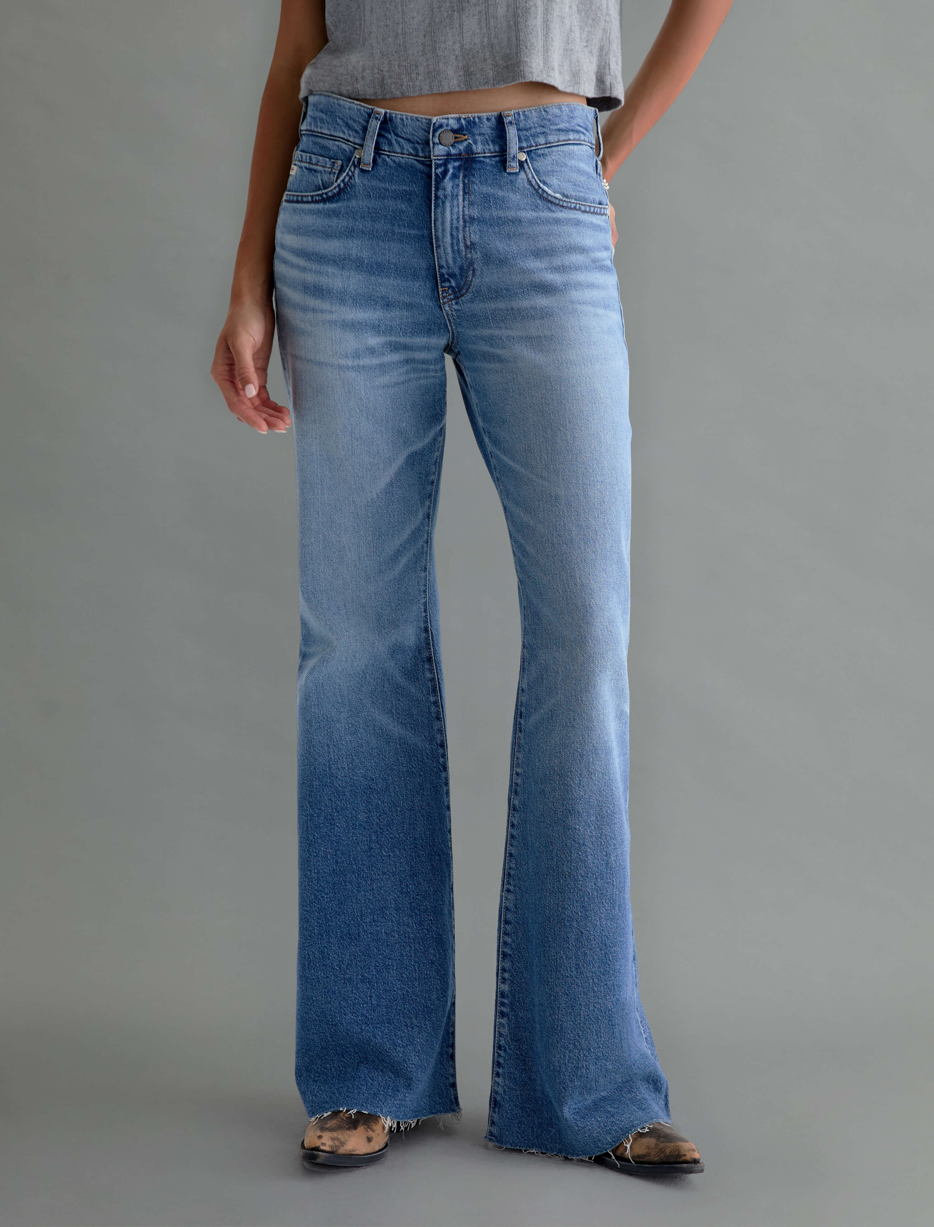 A person wearing light-to-medium blue wide-leg flare jeans, styled with a gray cropped top and brown flat sandals, standing against a solid gray studio background.