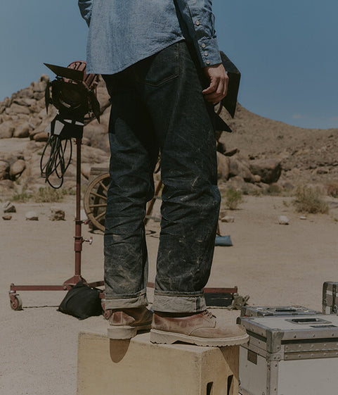 Person standing on a box in a desert setting with equipment and mountains in the background