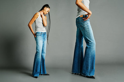 Model wearing blue flared jeans and white tank tops standing against a plain gray studio background