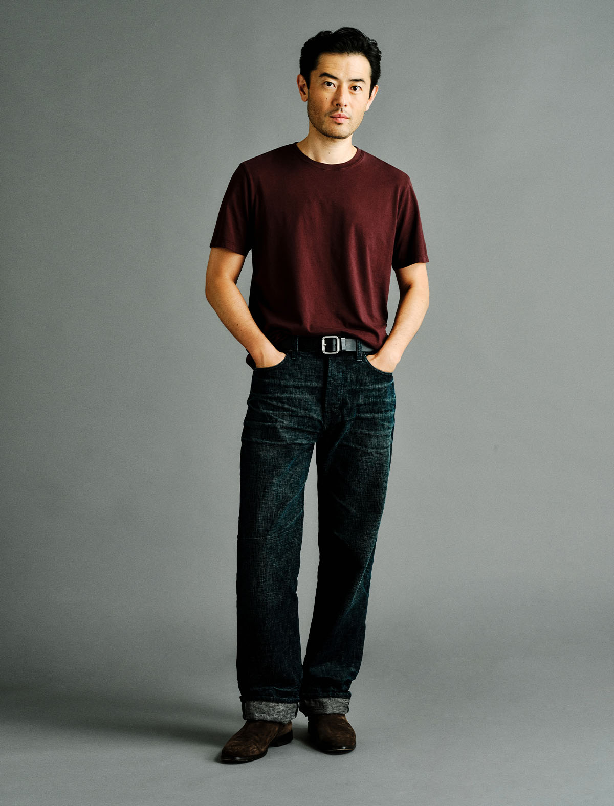 A man stands against a plain gray studio background wearing a dark burgundy short-sleeve t-shirt, dark blue jeans, a black belt, and brown shoes.