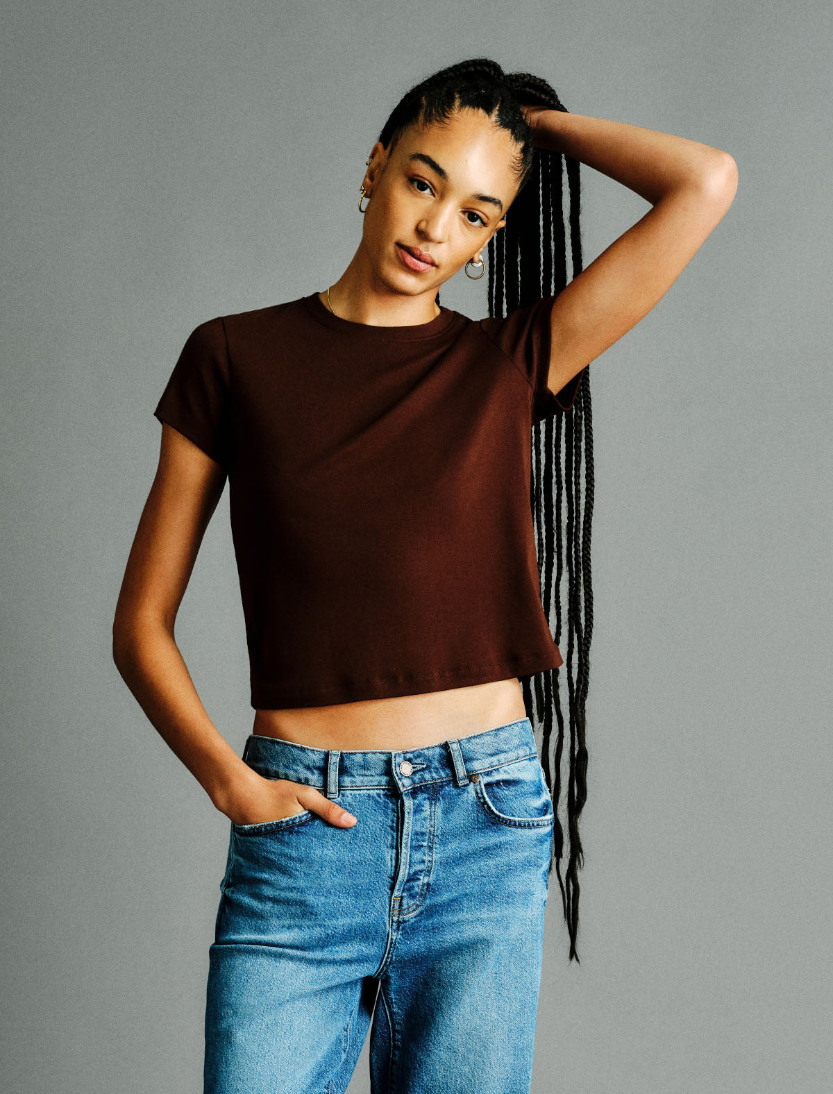 A woman modeling a dark brown short-sleeve crop top with blue jeans, posing against a solid gray studio background.