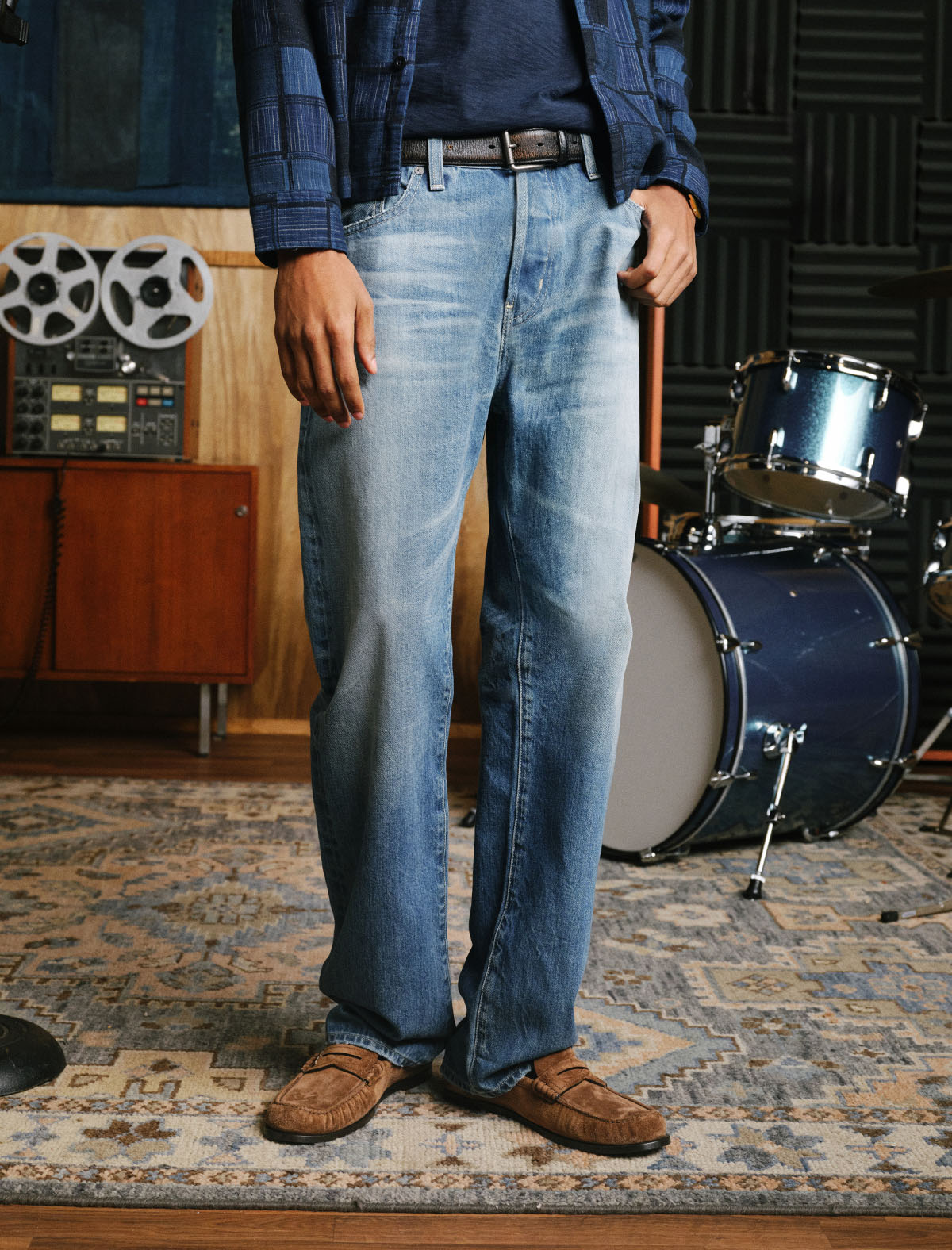 A person wearing light-wash blue jeans, a dark blue shirt, a blue plaid button-up shirt, and brown suede loafers, standing in a music studio with a drum set and reel-to-reel tape recorder in the background.