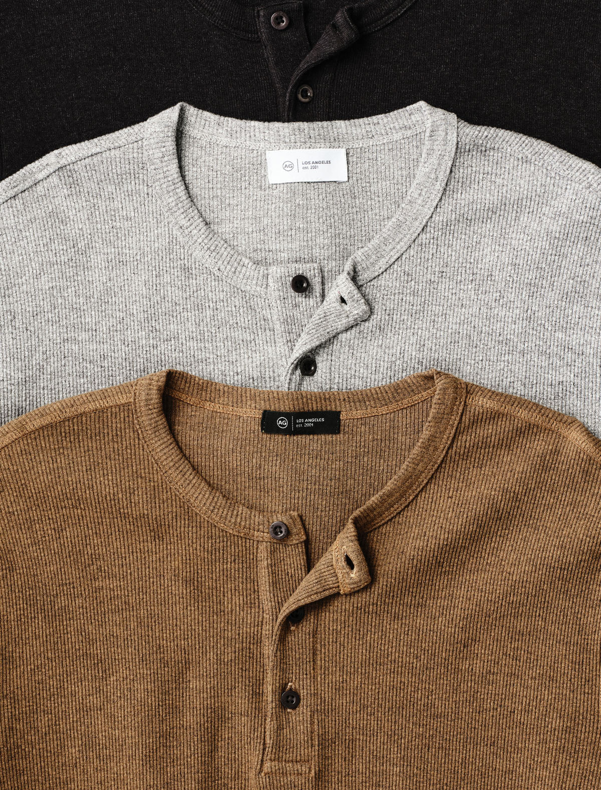 Three ribbed henley shirts stacked diagonally, showing the inner neck labels: a black shirt at the top, a light gray shirt in the middle, and a brown/tan shirt at the bottom.