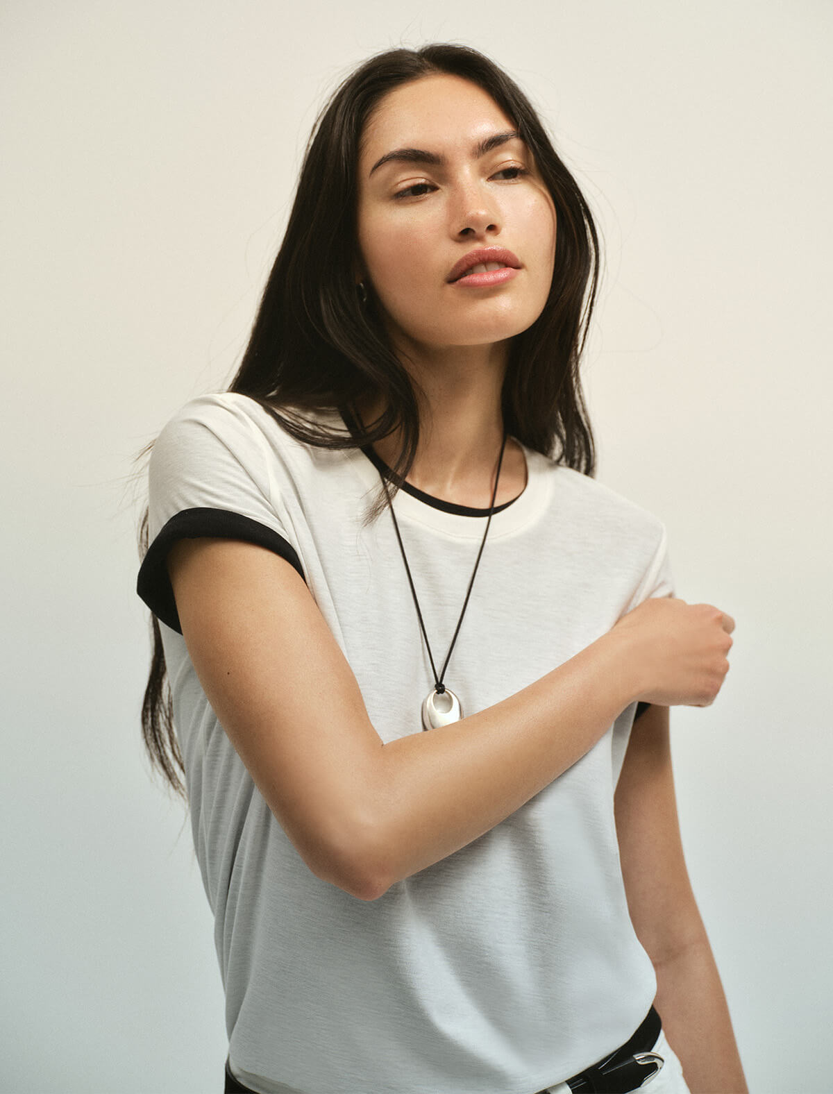 A woman wearing a light gray ringer t-shirt with black trim on the sleeves and neckline, styled with a silver pendant necklace, set against a plain light gray background.