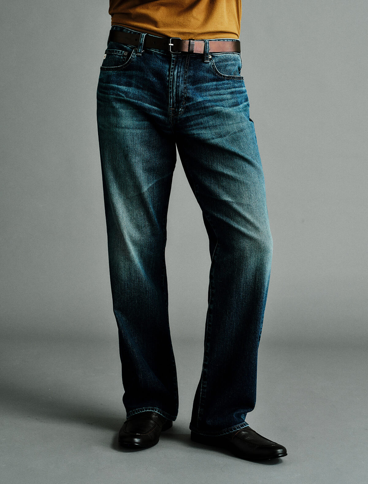 A person wearing dark blue straight-leg denim jeans with a brown leather belt, paired with a mustard yellow shirt and black leather shoes, standing against a neutral gray studio background.