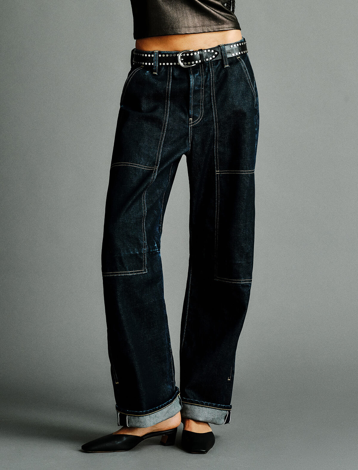 A person wearing dark indigo denim jeans with contrast light stitching, a black studded belt, and black pointed flats against a plain gray studio background.