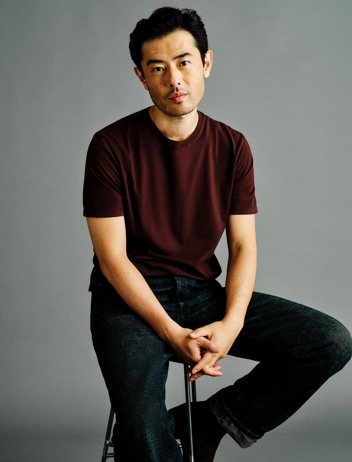 A man wearing a dark maroon crewneck t-shirt and dark blue jeans sits on a stool against a solid gray studio background.