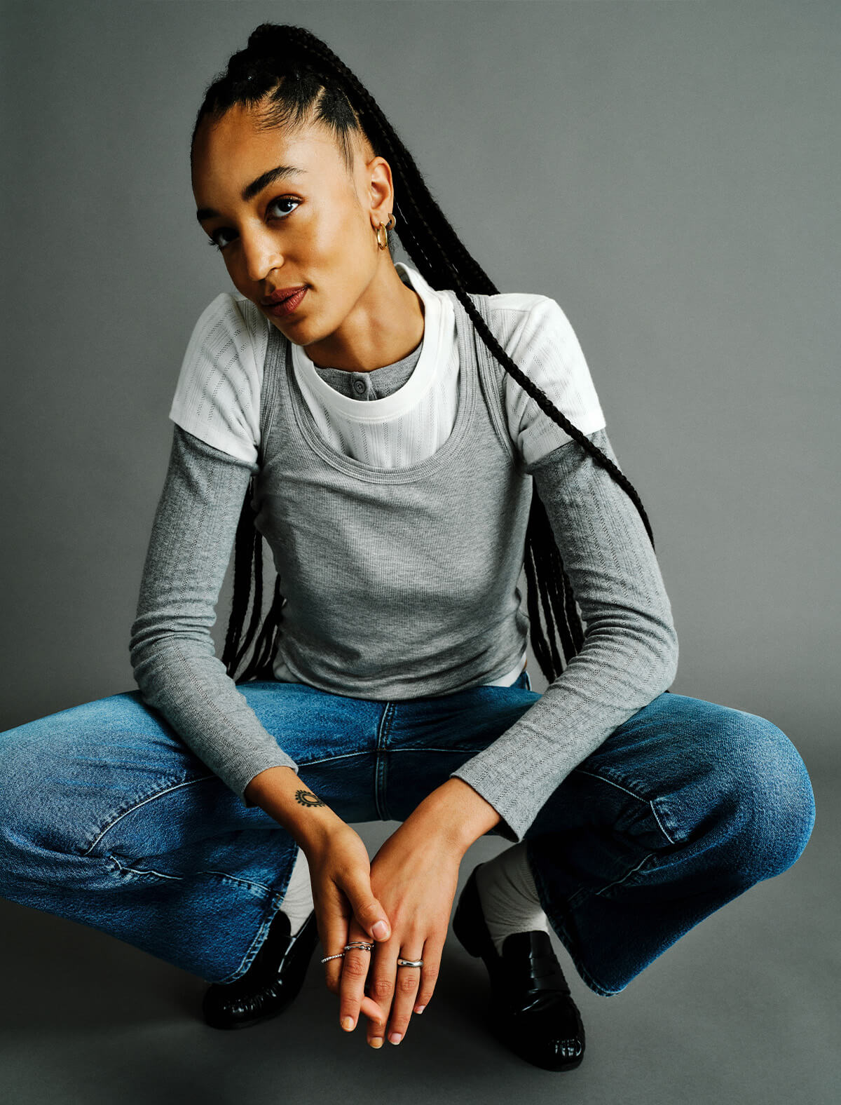 A model crouching in a studio setting, wearing a layered gray and white long-sleeve top, blue jeans, and black shoes against a solid gray background.