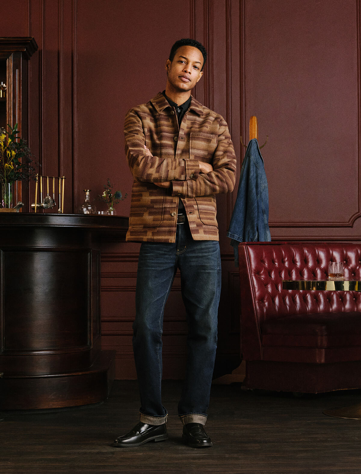 A man stands in a dimly lit, elegant room with dark wood paneling, wearing a brown and tan striped button-up shirt, dark blue jeans, and black dress shoes. A blue jacket hangs on a coat rack in the background next to a red tufted leather sofa.
