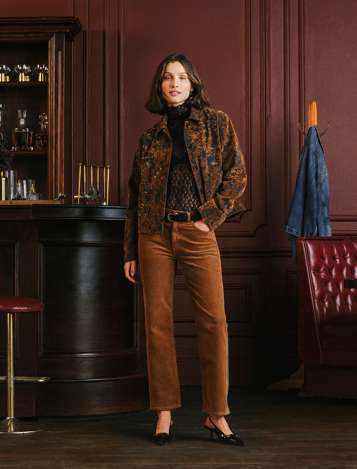 A woman stands in a dimly lit bar or lounge setting wearing a brown patterned jacket, black turtleneck, brown corduroy trousers, and black shoes. A red leather chair and dark wood paneling are visible in the background.