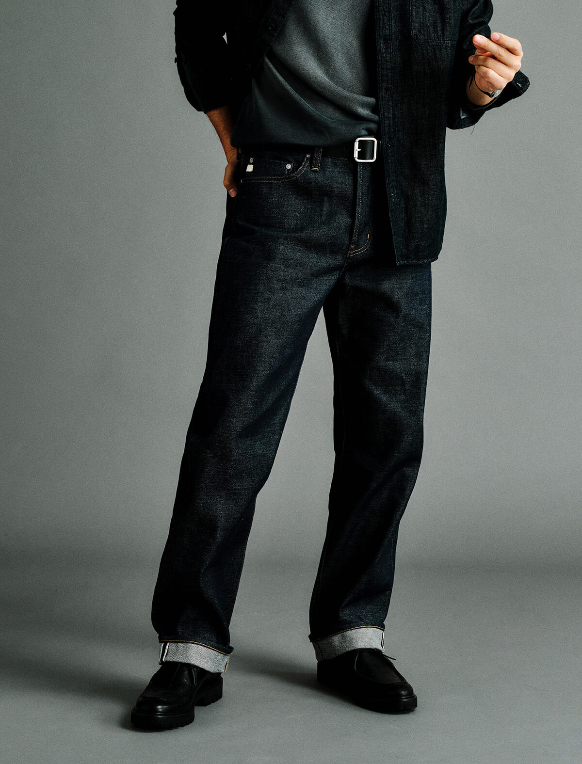 A person wearing dark indigo denim jeans with a black belt, styled with a dark shirt, jacket, and black shoes against a plain gray studio background.