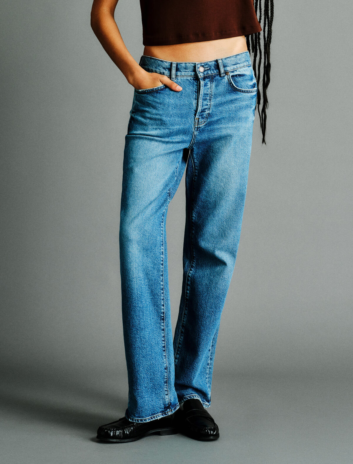 A person wearing medium-wash blue straight-leg jeans, a dark brown cropped top, and black shoes, standing against a plain gray studio background.