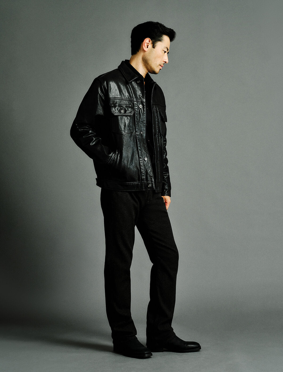 A man wearing a black leather jacket, black shirt, black trousers, and black shoes, standing in profile against a solid gray studio background.