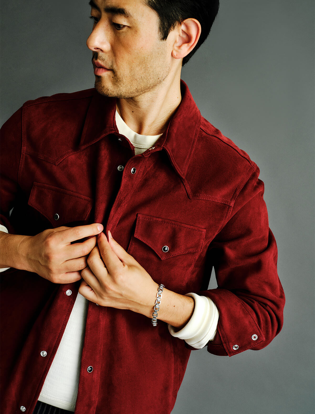 A man wearing a dark red corduroy button-up shirt jacket over a white crewneck t-shirt, adjusting the collar with one hand and buttoning the front with the other. He is wearing a silver chain bracelet on his wrist.