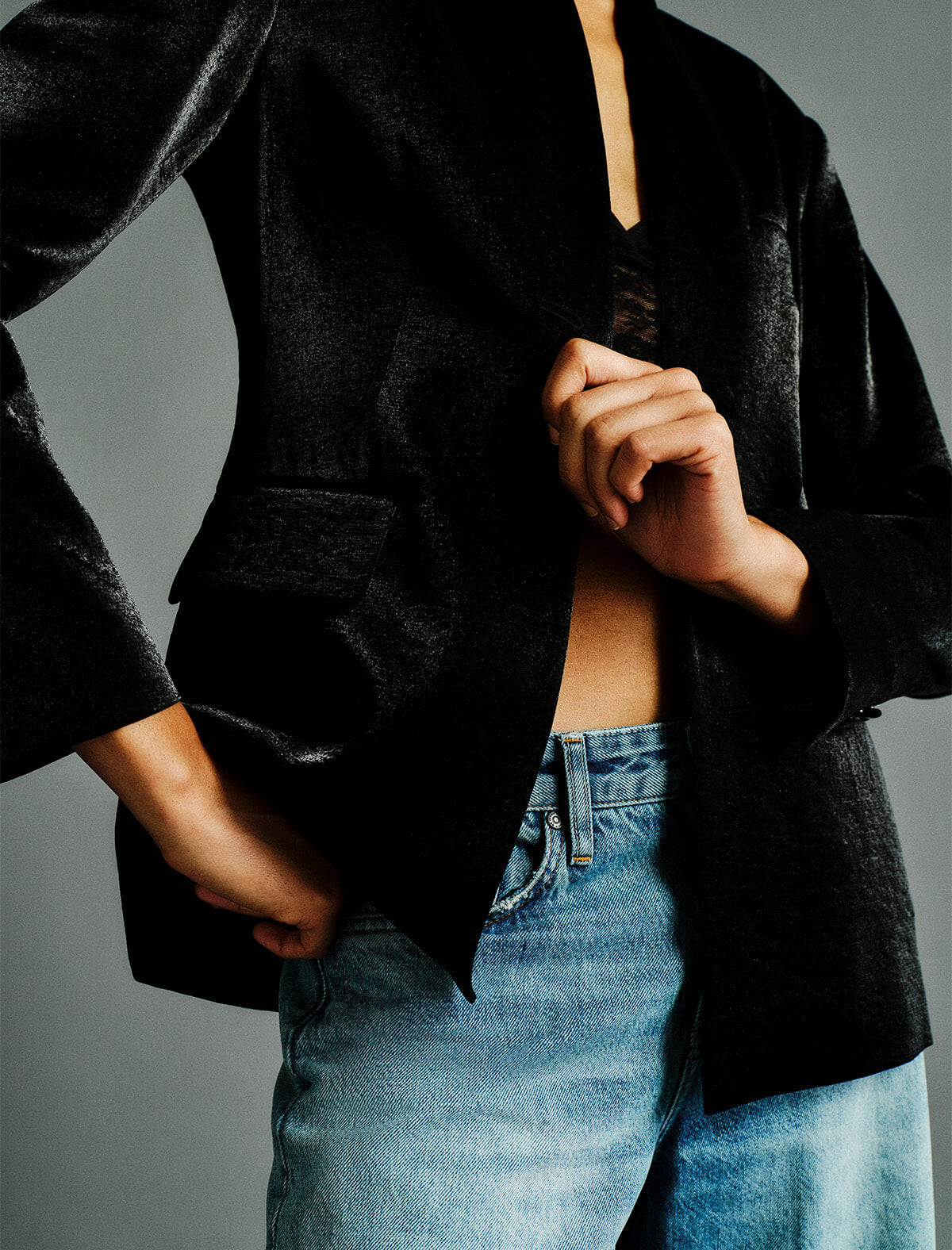 A close-up of a person wearing an oversized black velvet blazer over a black lace top and light blue jeans, set against a neutral gray studio background.