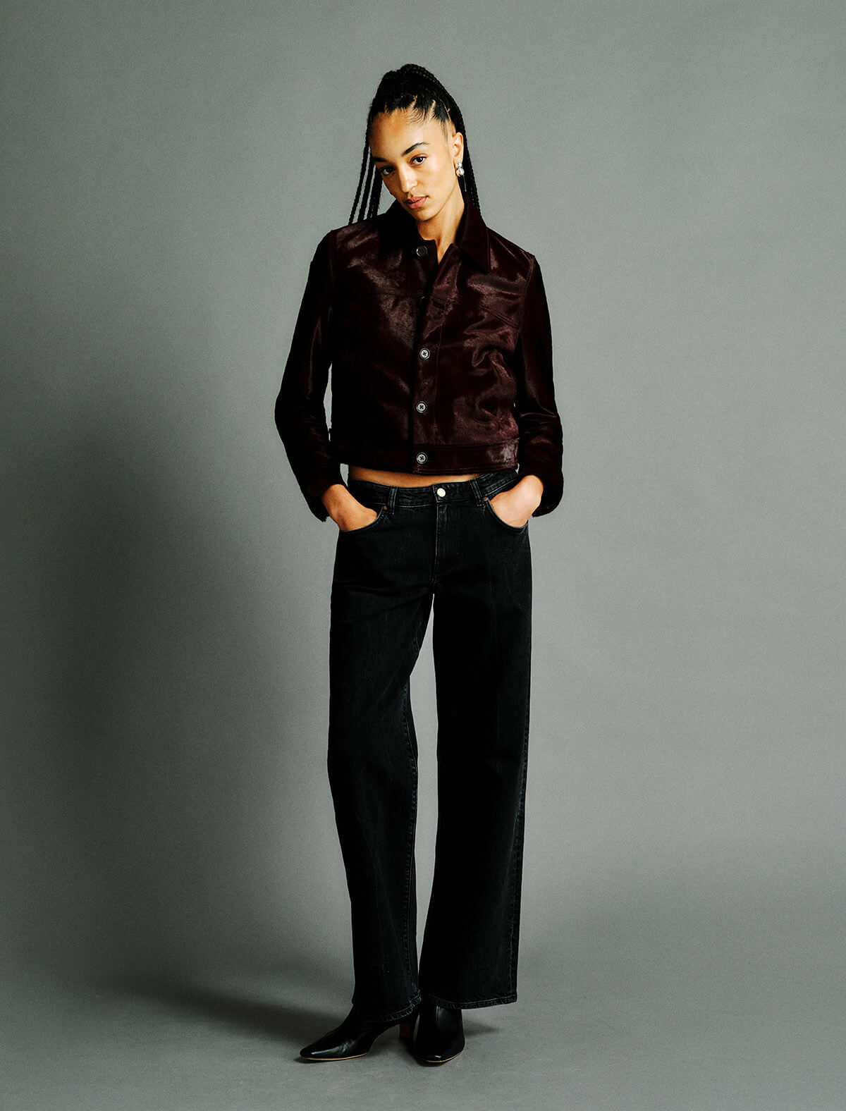 A model stands against a solid gray studio background wearing a dark brown satin button-up shirt and black wide-leg jeans.