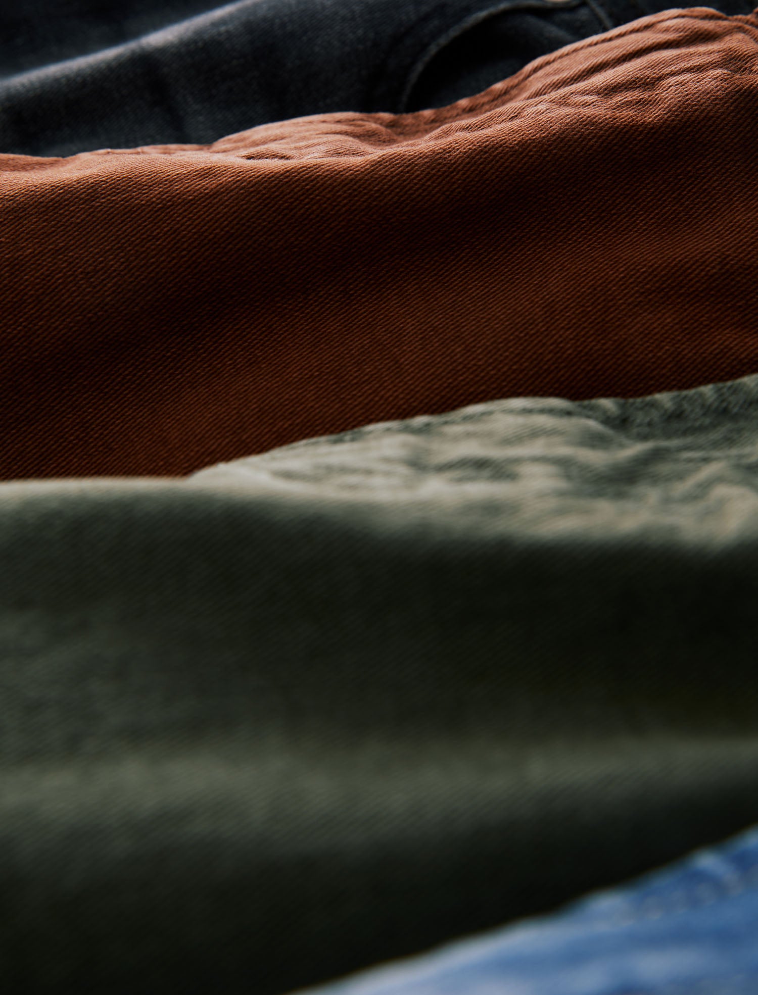 A close-up, angled view of folded fabric layers in earthy tones, featuring a prominent rust-brown textile, a dark olive-green textile, and a dark blue textile.