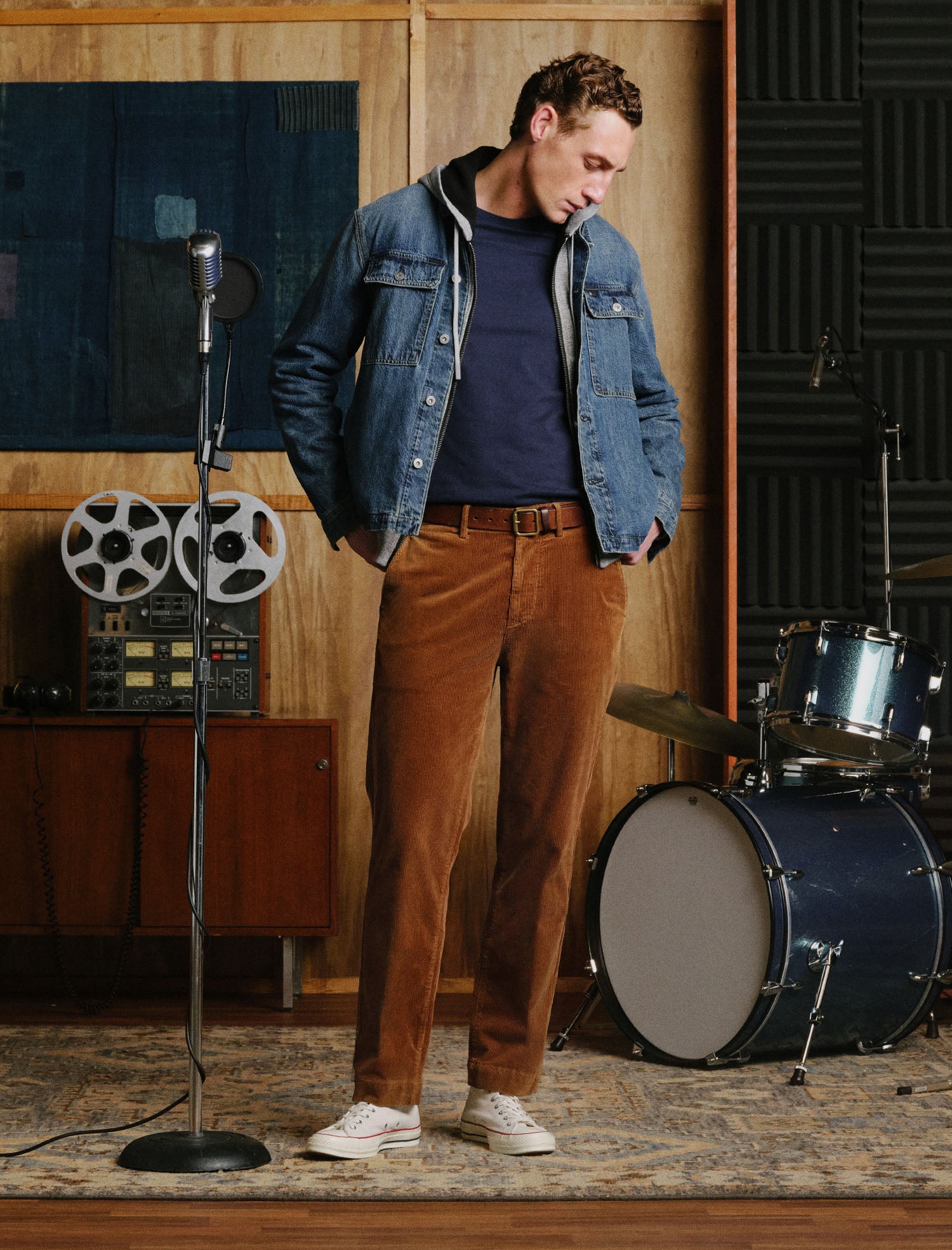A man stands in a music studio wearing a blue denim jacket over a navy t-shirt, brown corduroy pants, and white sneakers. A vintage microphone, reel-to-reel tape machine, and drum set are visible in the background.