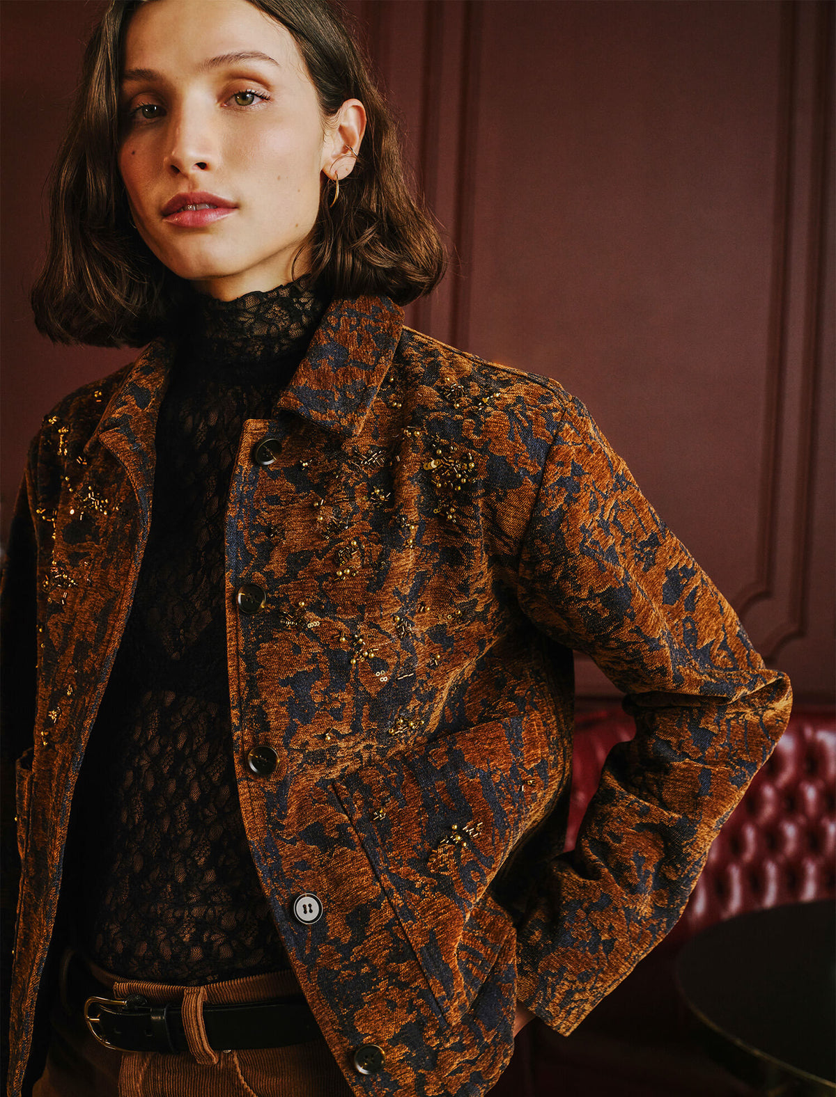 A woman wearing a brown and black patterned textured jacket over a black lace turtleneck, photographed in an indoor setting with a dark red wall and tufted chair in the background.