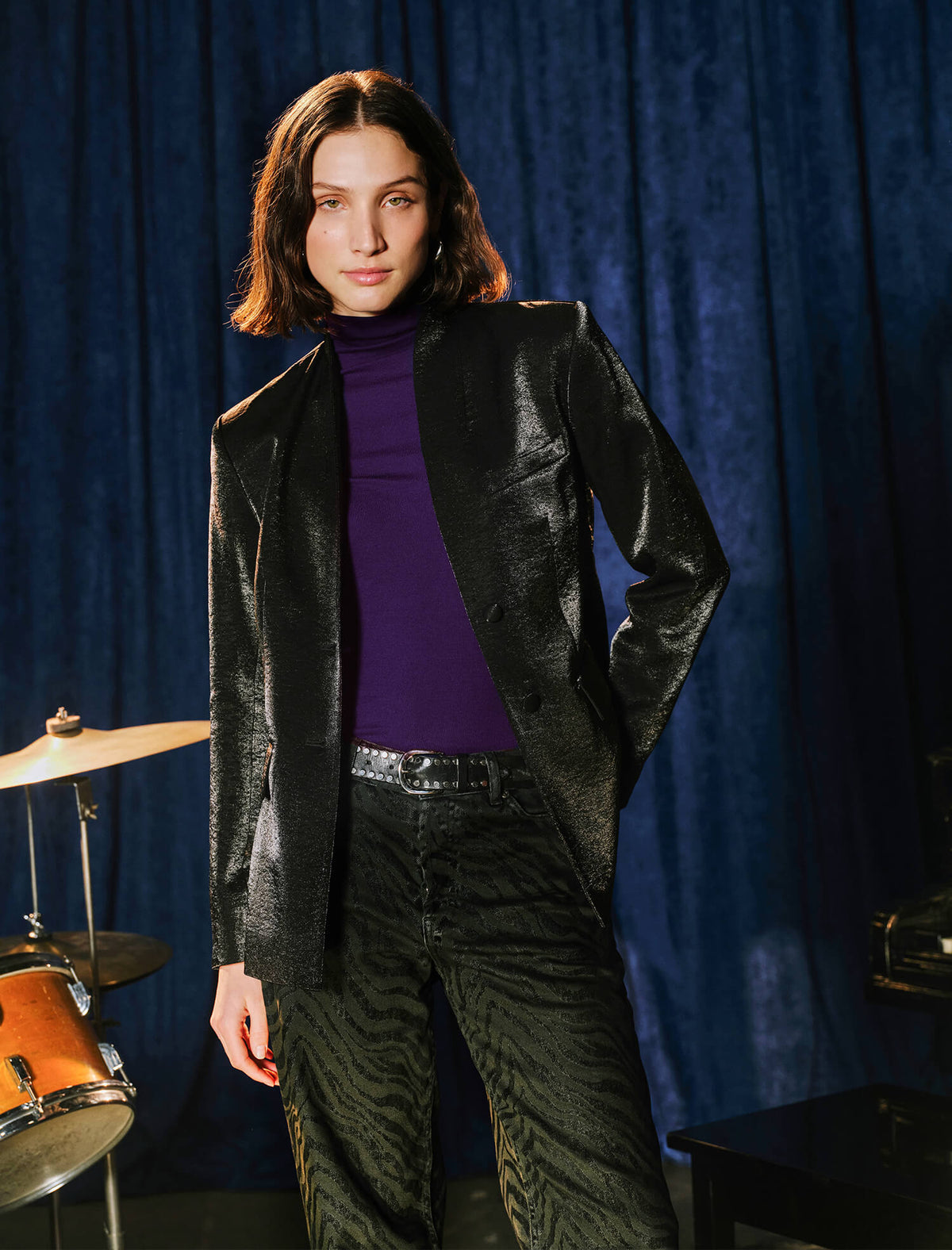 A model stands in front of a dark blue velvet curtain wearing a black textured blazer over a purple turtleneck, paired with dark patterned trousers. A cymbal and other studio equipment are partially visible in the background.