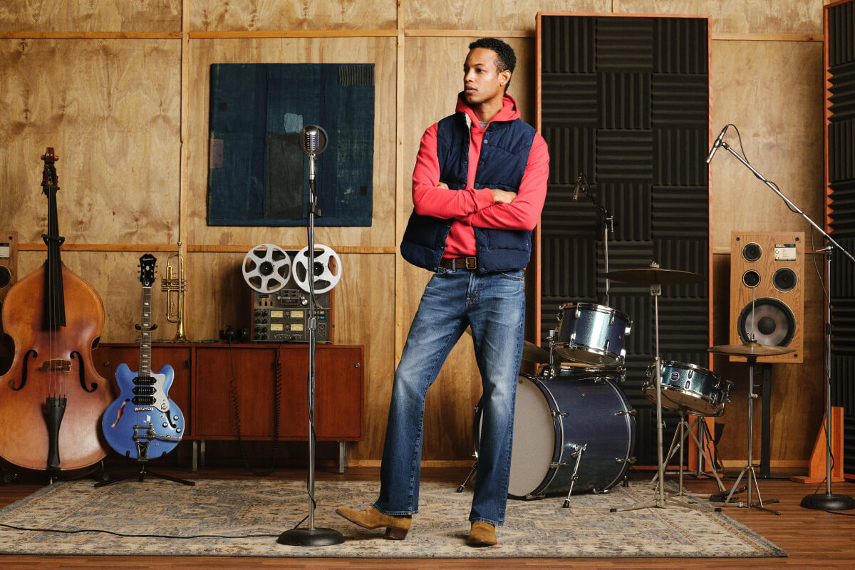 A man stands in a music studio wearing a bright pink long-sleeve shirt under a navy blue vest, blue jeans, and brown boots. He is posed with arms crossed in front of a microphone stand, with a double bass, blue electric guitar, film reels, drums, and speakers in the background.