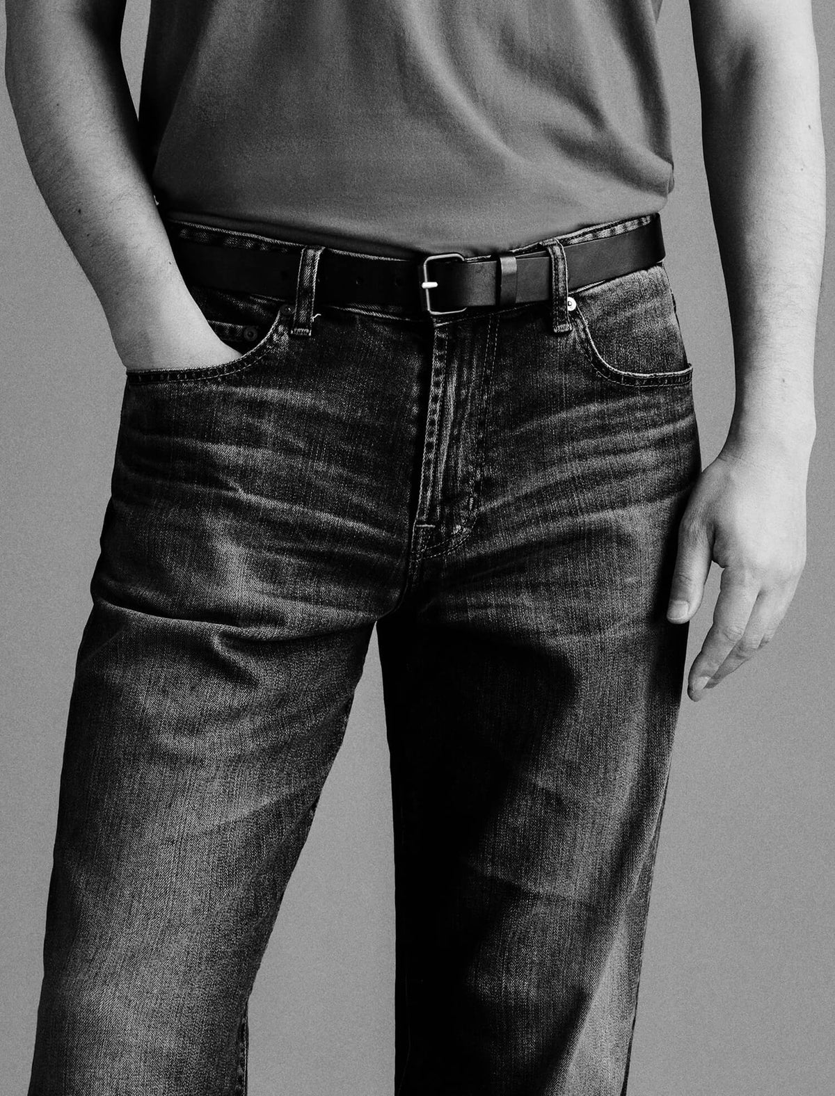 A close-up of a person wearing dark-wash denim jeans with a black belt, paired with a gray t-shirt, shown in a black and white photograph against a neutral gray background.