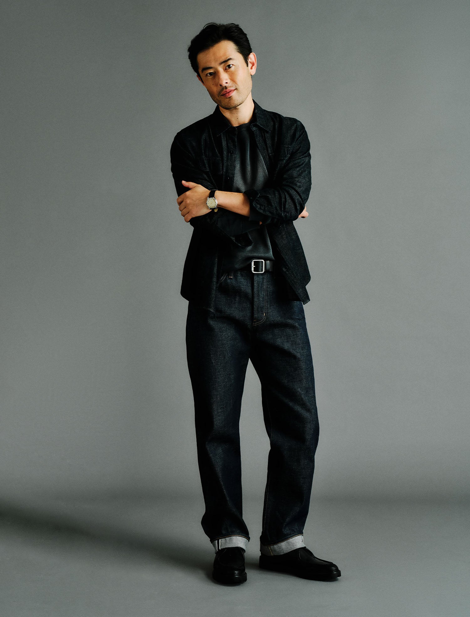 A man wearing a black long-sleeve button-up shirt, dark blue jeans, and black shoes, standing with arms crossed against a solid gray studio background.