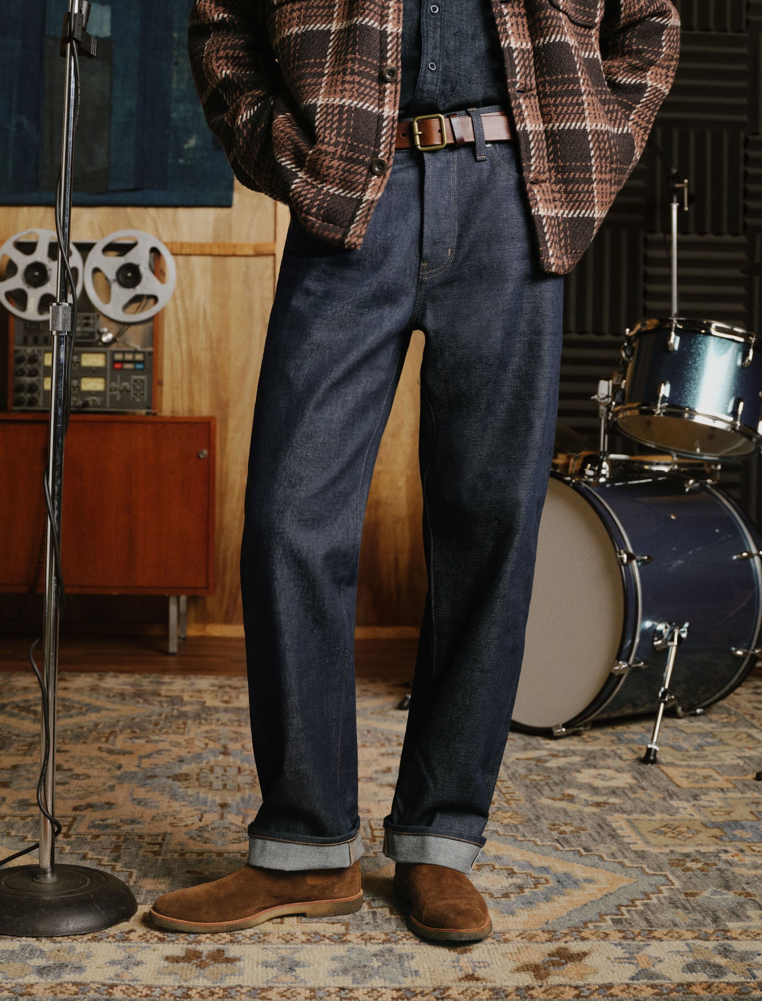 A person standing indoors wearing dark blue denim jeans with rolled cuffs, a brown plaid overshirt, a dark shirt, and brown suede shoes. The setting includes a patterned rug, wood paneling, a reel-to-reel machine, and a drum set in the background.