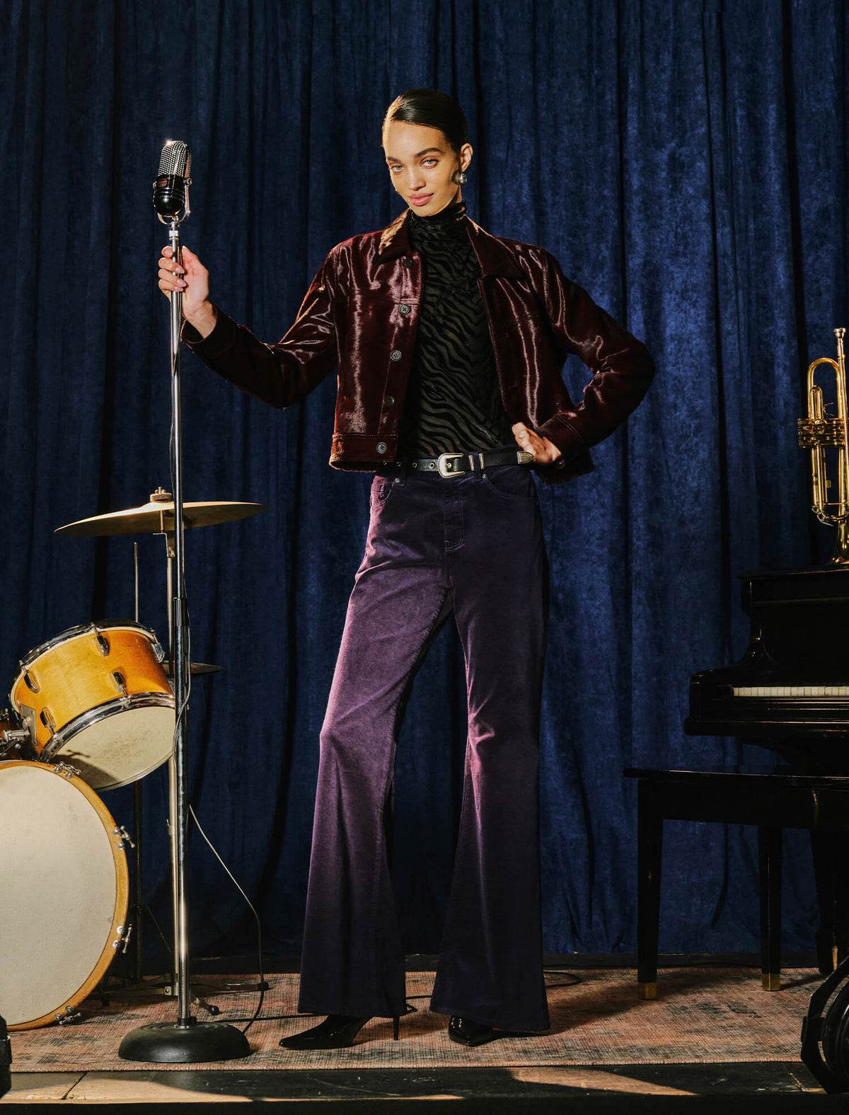 A model stands on a stage-like set in front of a blue velvet curtain, holding a vintage microphone stand. The outfit features a deep burgundy velvet jacket, a black-and-white patterned turtleneck, and purple wide-leg velvet pants.