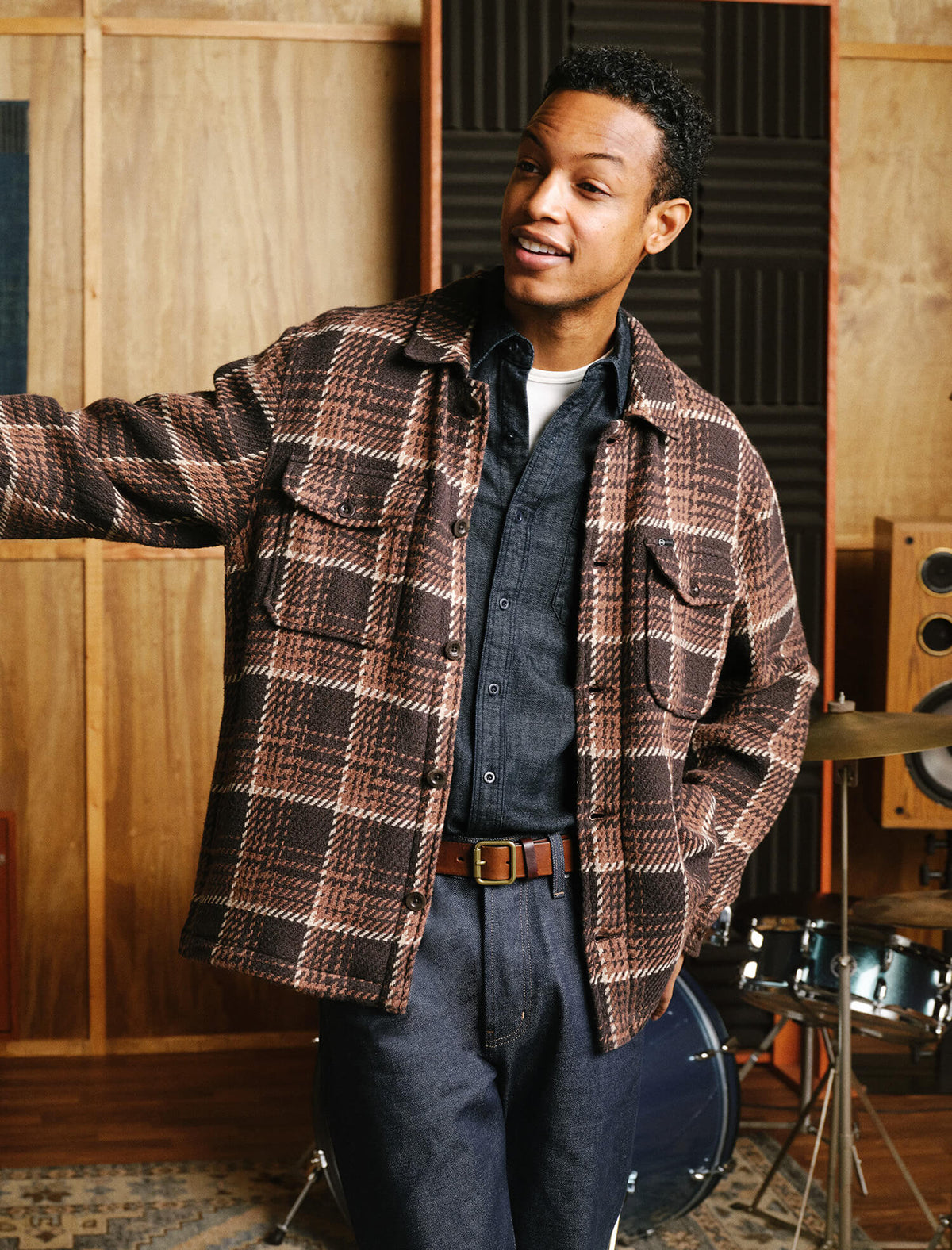 A man wearing a brown and black plaid overshirt over a dark blue button-up shirt and jeans, standing in a wood-paneled room with musical equipment in the background.