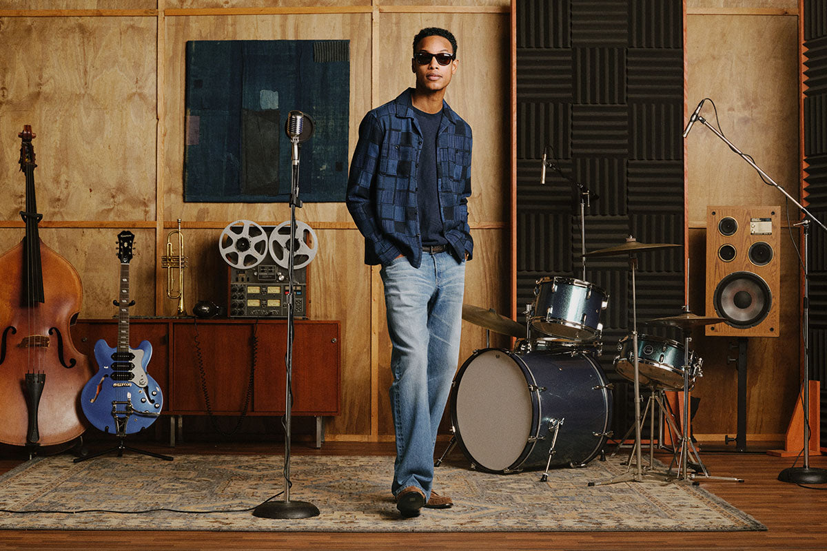 A man wearing sunglasses, a blue plaid button-down shirt, and light-wash jeans stands in a music studio with guitars, a drum set, microphones, and wood-paneled walls.