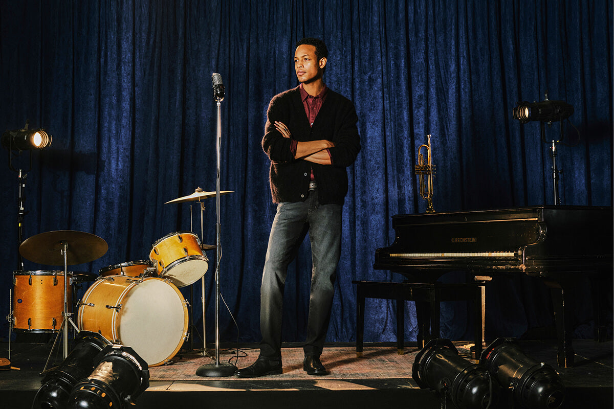 A man stands on a stage in front of a blue velvet curtain with a microphone, drums, and a grand piano around him. He is wearing a dark cardigan over a red shirt and blue jeans.