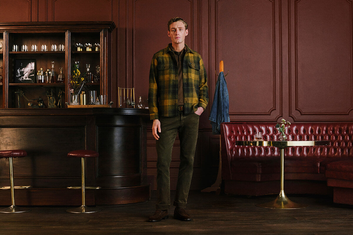 A man stands in a dimly lit bar interior wearing a green and yellow plaid flannel shirt, olive green trousers, and brown boots. A dark wood bar, red leather booth, and wood-paneled walls are visible in the background.