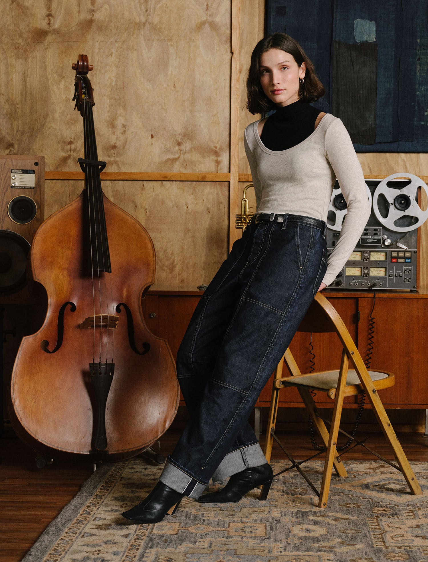 A woman leans against a yellow folding chair in a rustic studio setting, wearing a beige long-sleeve top, dark blue wide-leg jeans, and black ankle boots. A large double bass stands to her left, with vintage audio equipment behind her.