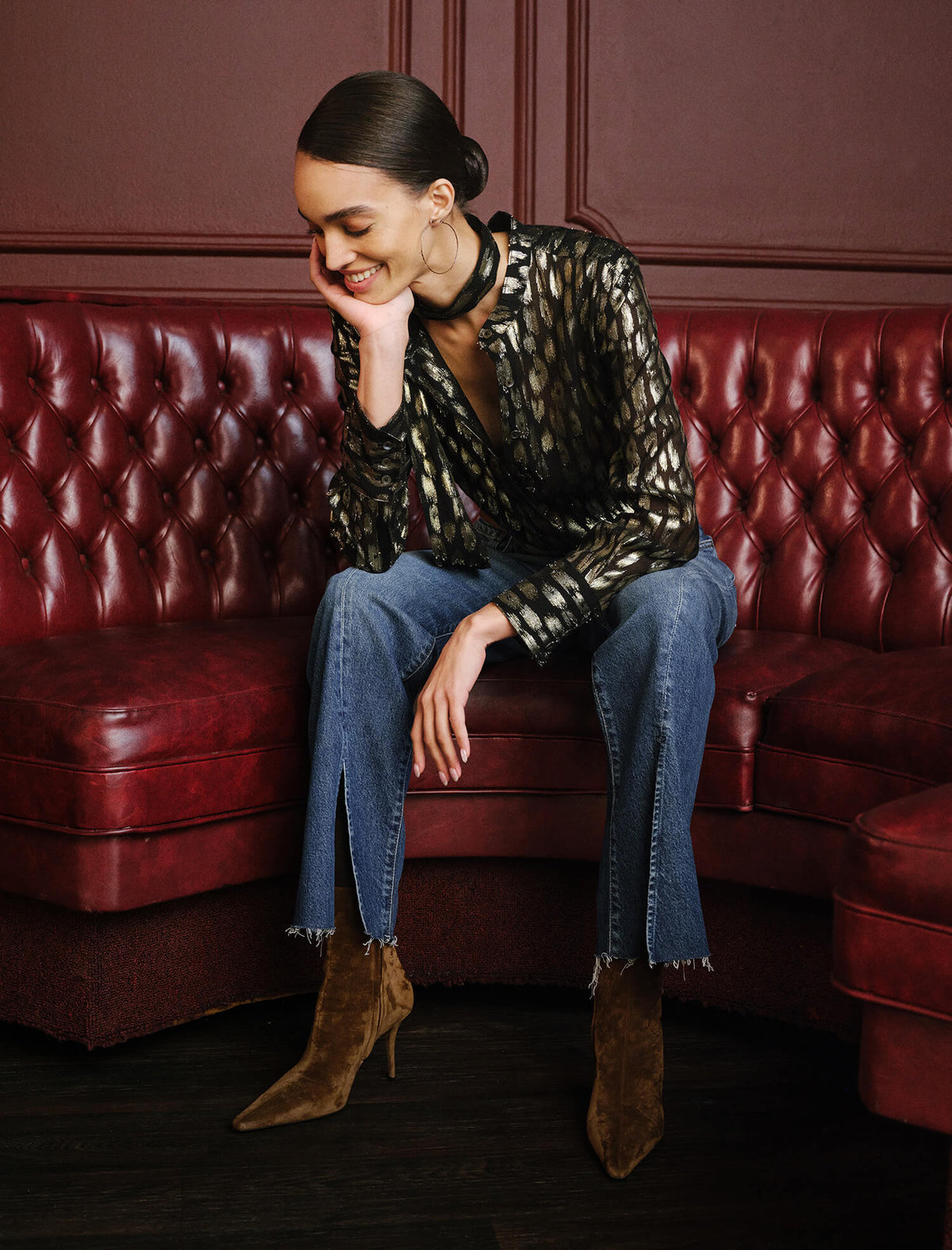 A woman sits on a tufted red leather sofa wearing a sheer black and gold patterned blouse, blue jeans, and brown suede ankle boots in a dark, elegant room.