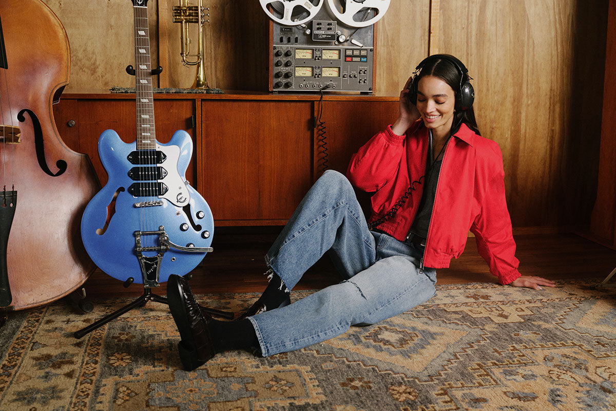 A woman wearing a red jacket, blue jeans, and black headphones sits on a patterned rug in a music room, leaning against a wooden wall next to a blue electric guitar, a cello, and a reel-to-reel tape recorder.