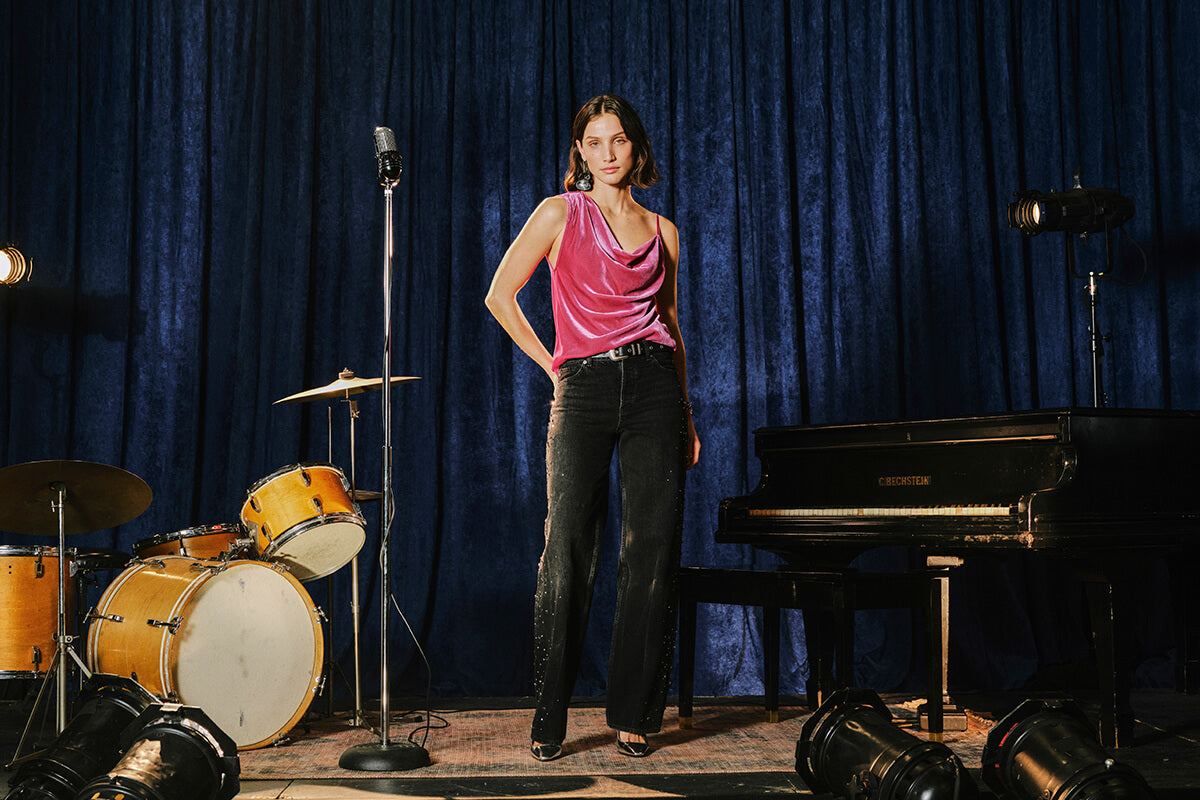 A woman stands on a stage-like set with a deep blue velvet curtain backdrop, wearing a sleeveless pink top and dark jeans. A drum set, microphone, piano, and stage lights are visible around her.