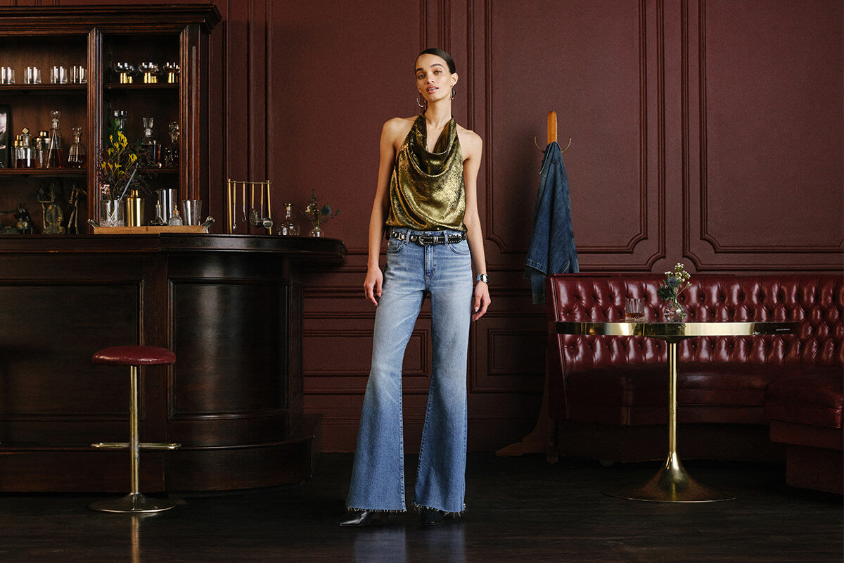 A model stands in a dimly lit, elegant bar interior wearing a metallic gold cowl-neck top and light-wash blue flared jeans.