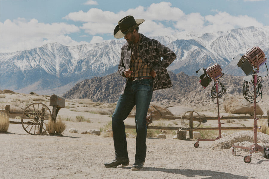 Person in cowboy hat and plaid shirt standing in a desert with mountains in the background