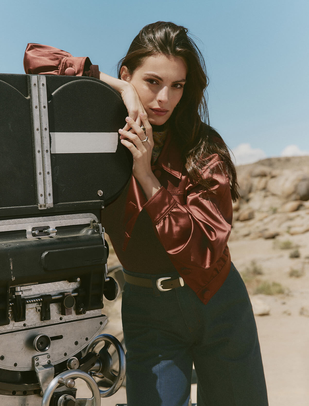 Woman in a red blouse and dark pants leaning against a large black case in a desert setting.