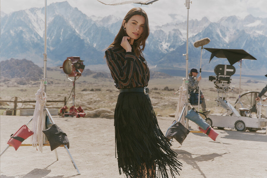 Woman in a black patterned top and fringed skirt on a film set with mountains in the background