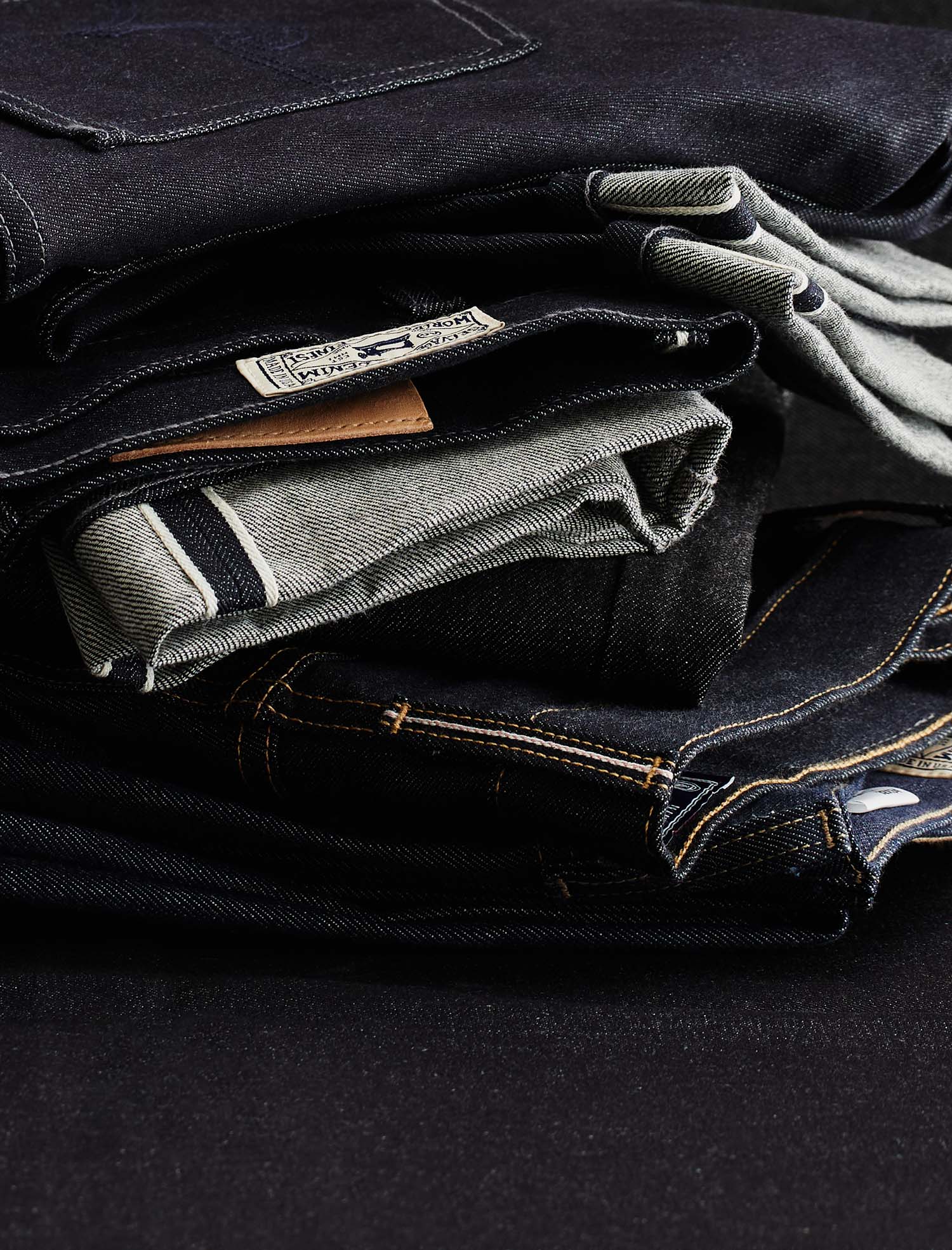 A close-up stack of folded dark denim jeans showing different washes and details like contrast stitching, pocket edges, and a fabric label.