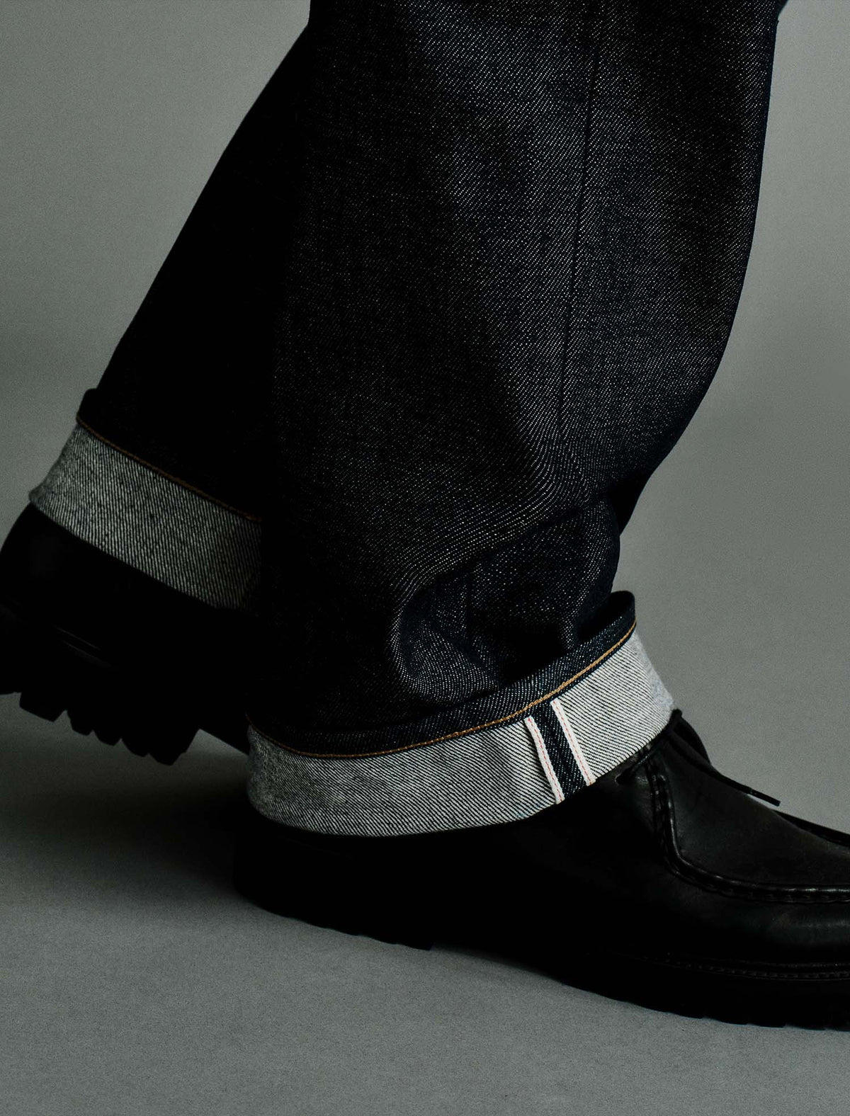 Close-up of dark denim jeans with a turned-up cuff showing a light gray inner lining and contrast stitching, paired with black leather shoes on a gray studio background.
