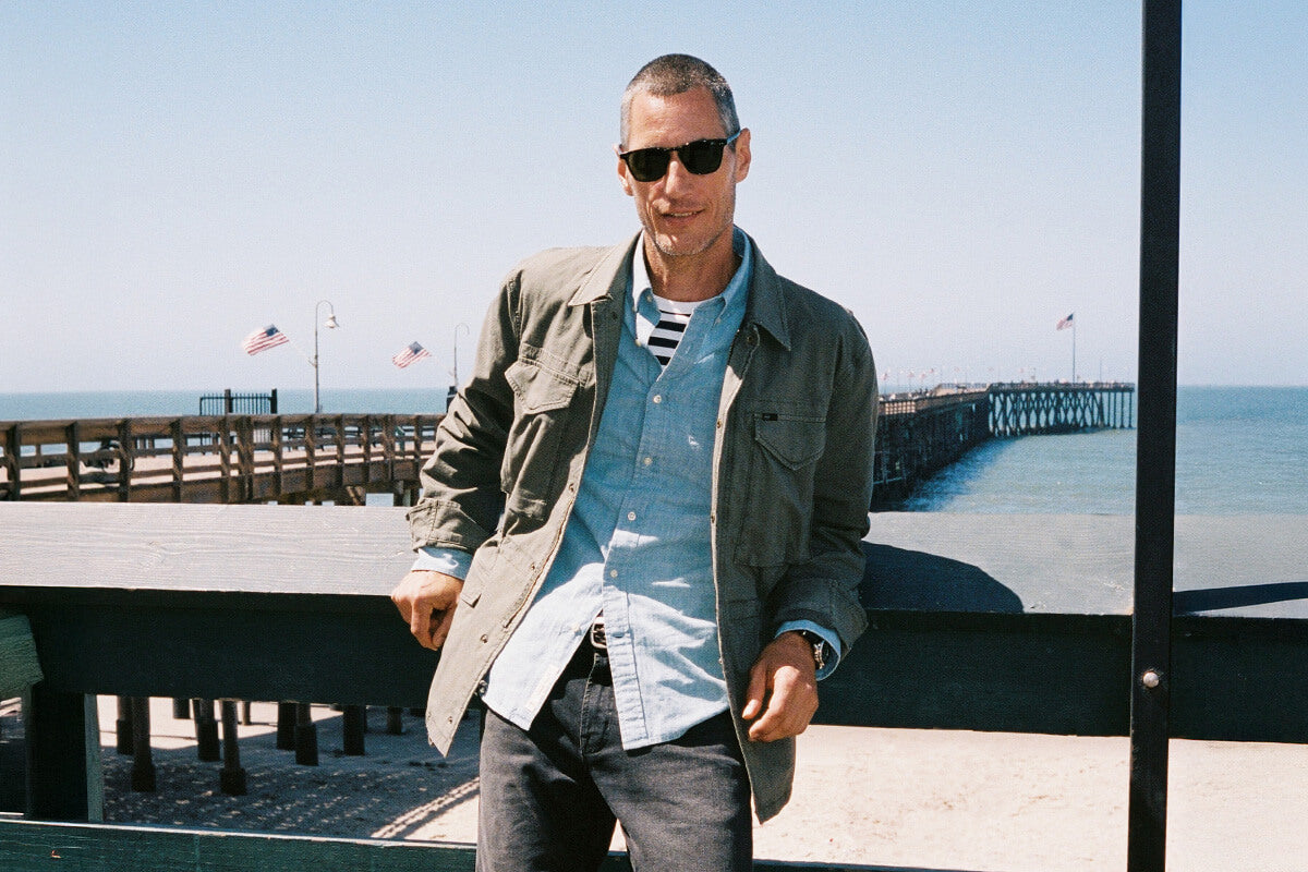 Man standing on a pier wearing sunglasses and casual clothing with a clear sky and water background.