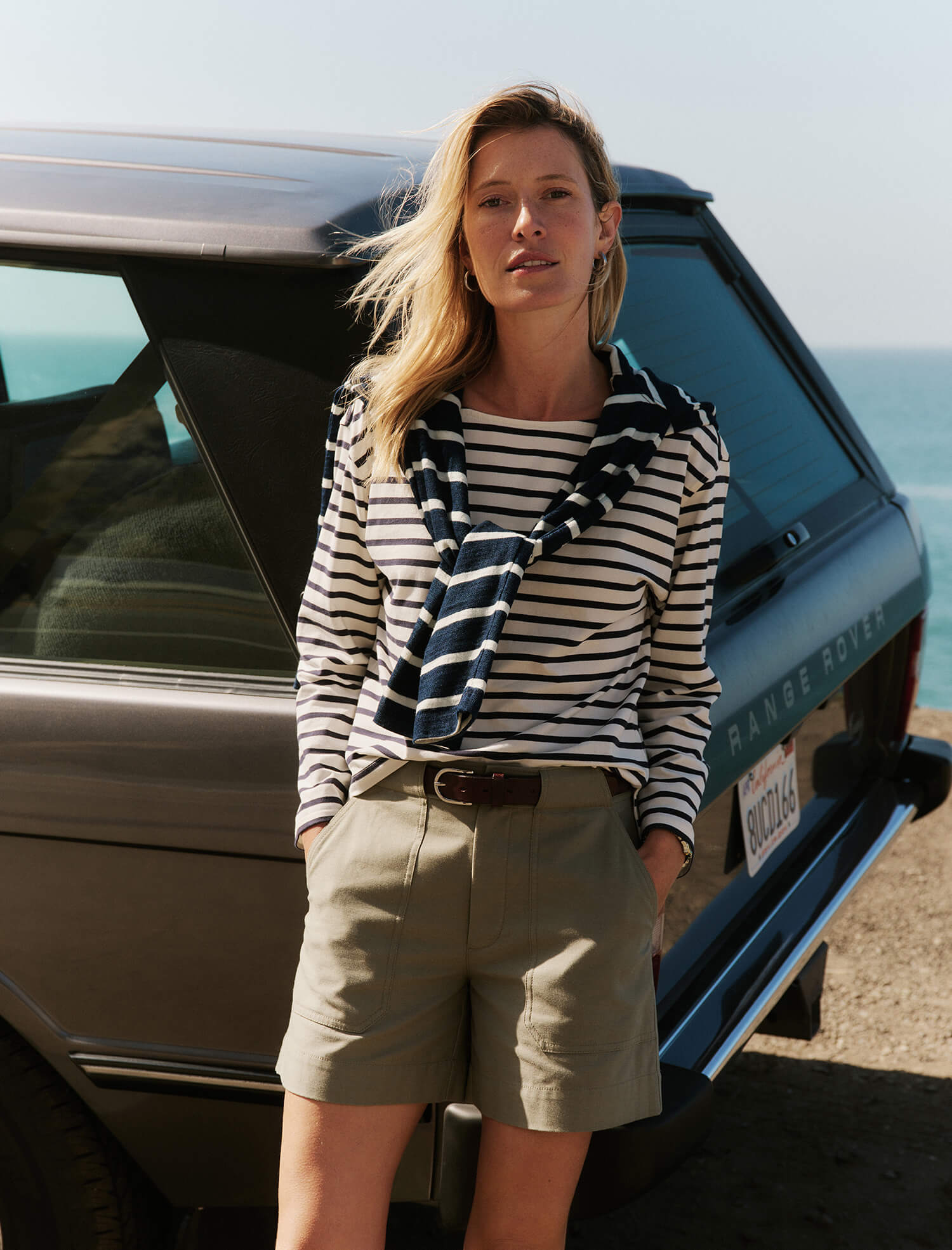 Woman standing next to a car by the sea, wearing a striped shirt and shorts.