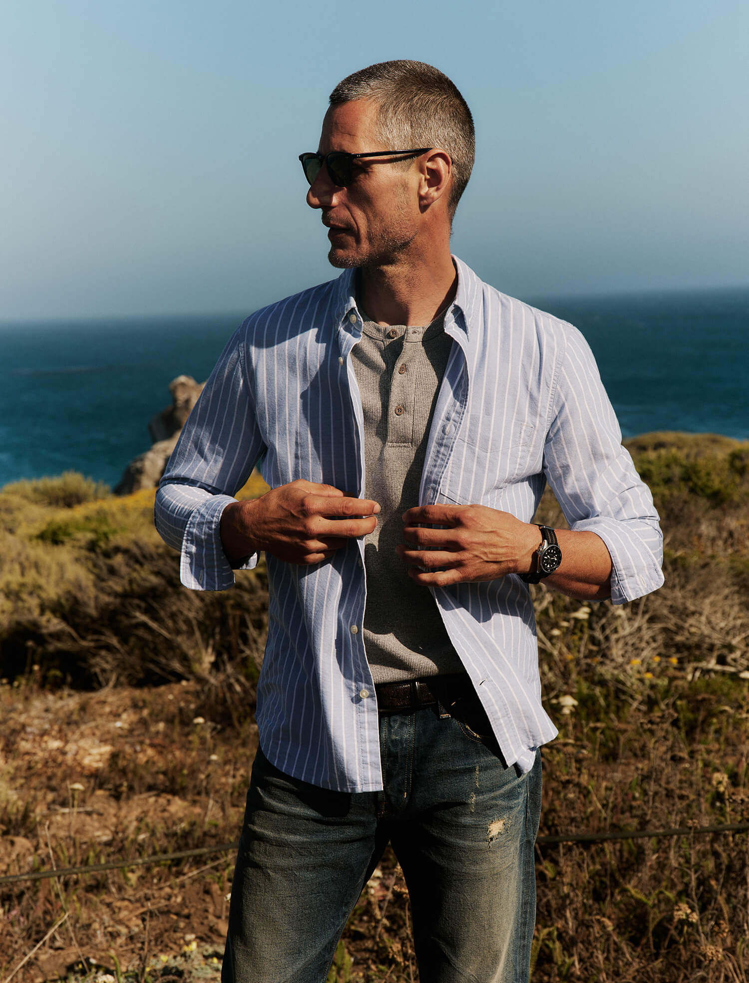 Man standing on a coastal cliffside wearing sunglasses and a striped shirt.