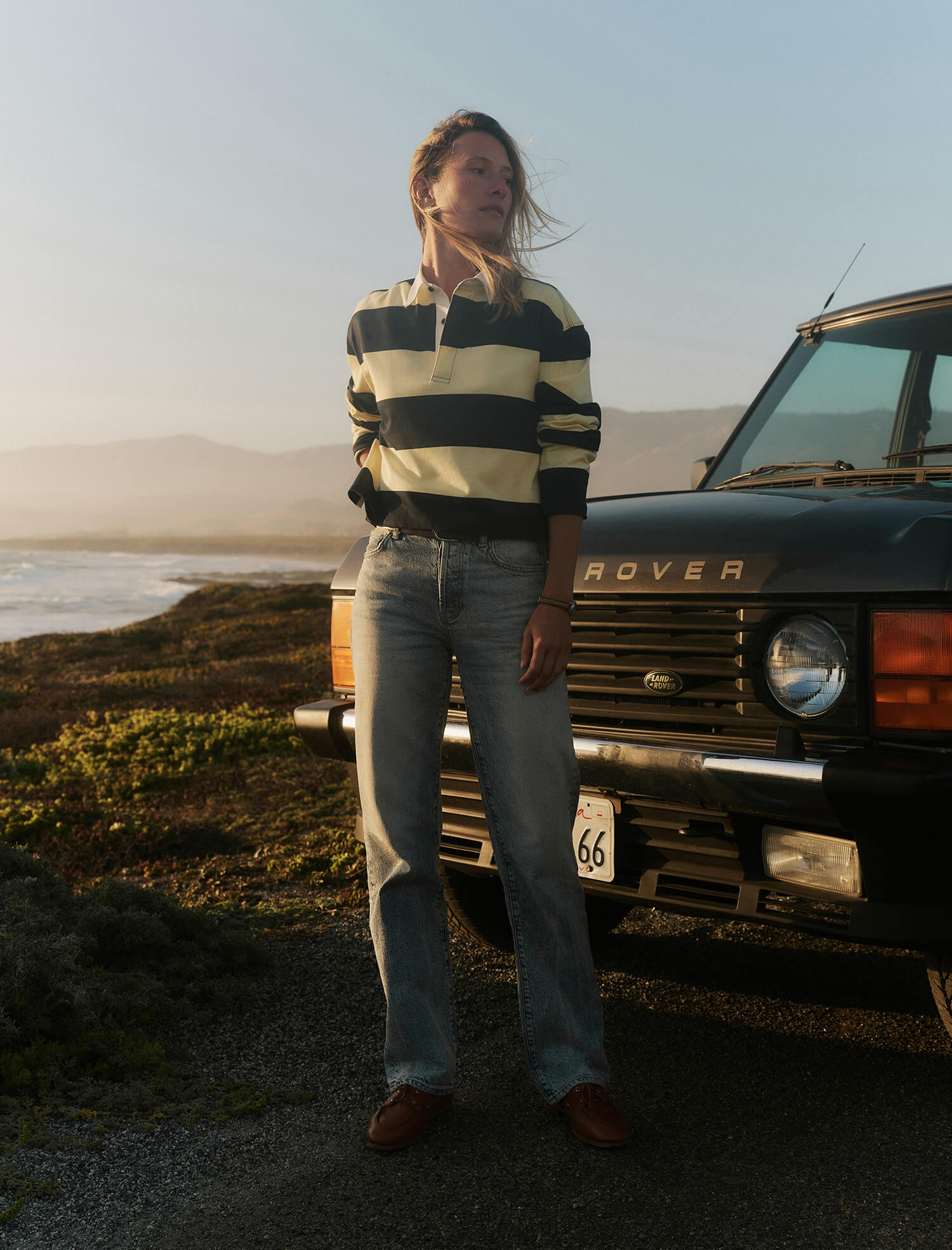 Person standing in front of a vintage Land Rover by a coastal road.