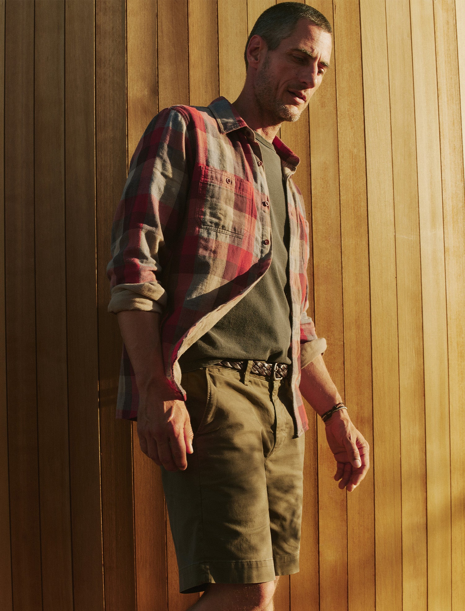 Man wearing a plaid shirt and shorts against a wooden wall